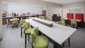 Un espace repas moderne avec une longue table blanche, des chaises vertes, un comptoir doté de différents distributeurs de nourriture et de boissons, et un mur d’accent rouge.