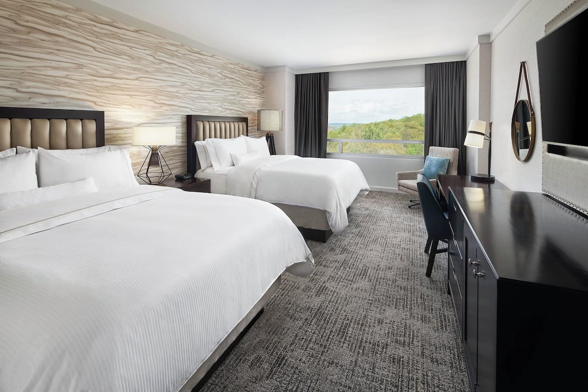 A hotel room with two beds, a desk, a chair, a TV, and a large window with a view.