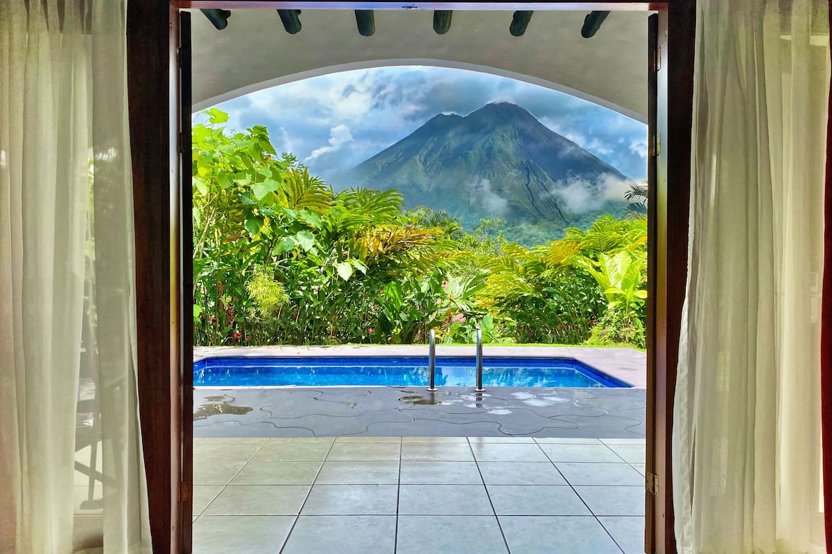 A view through an arched doorway to a swimming pool surrounded by lush greenery and a mountain in the background.