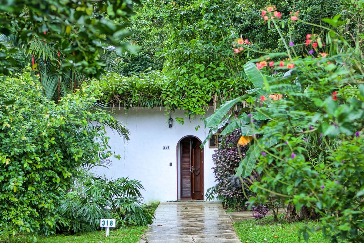 A white building with a wooden door and a small porch area surrounded by lush greenery and flowering plants.