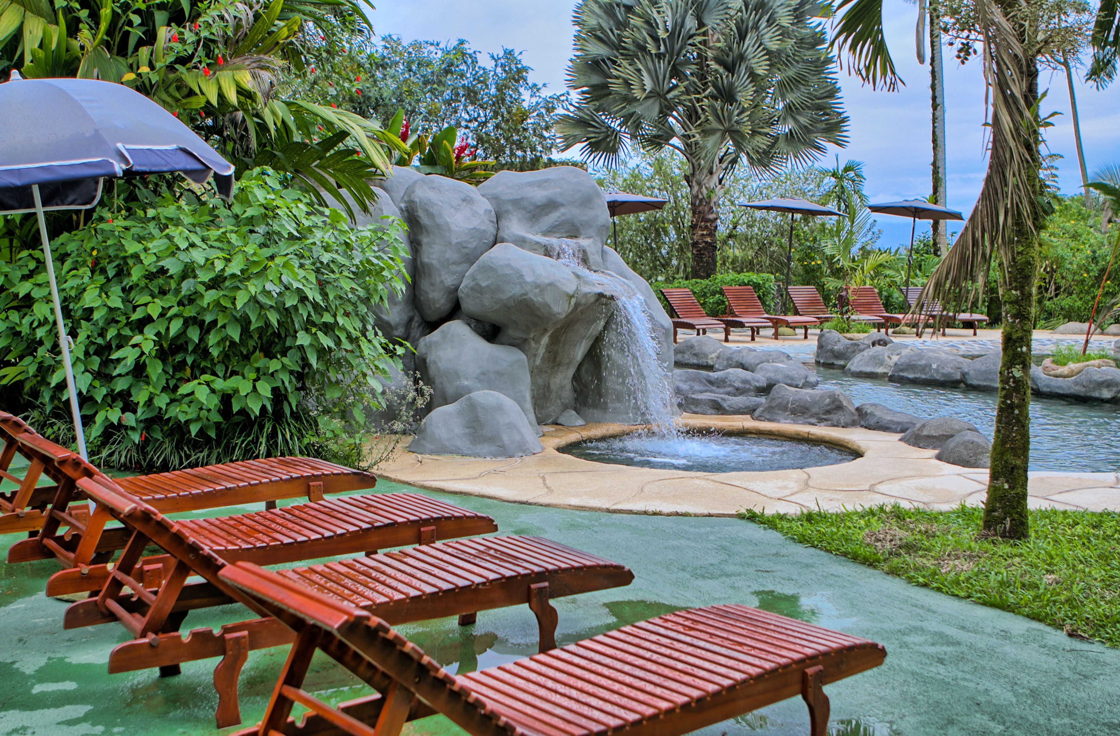 Uma área de piscina com espreguiçadeiras de madeira, uma cascata e palmeiras.