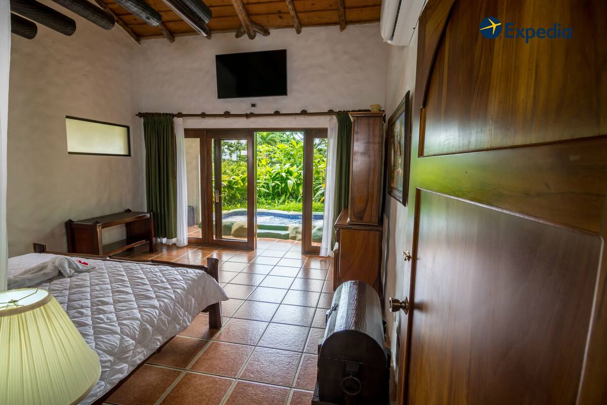 A bedroom with a bed, a bedside table, a lamp, a wooden wardrobe, and a sliding glass door leading to a balcony with greenery.