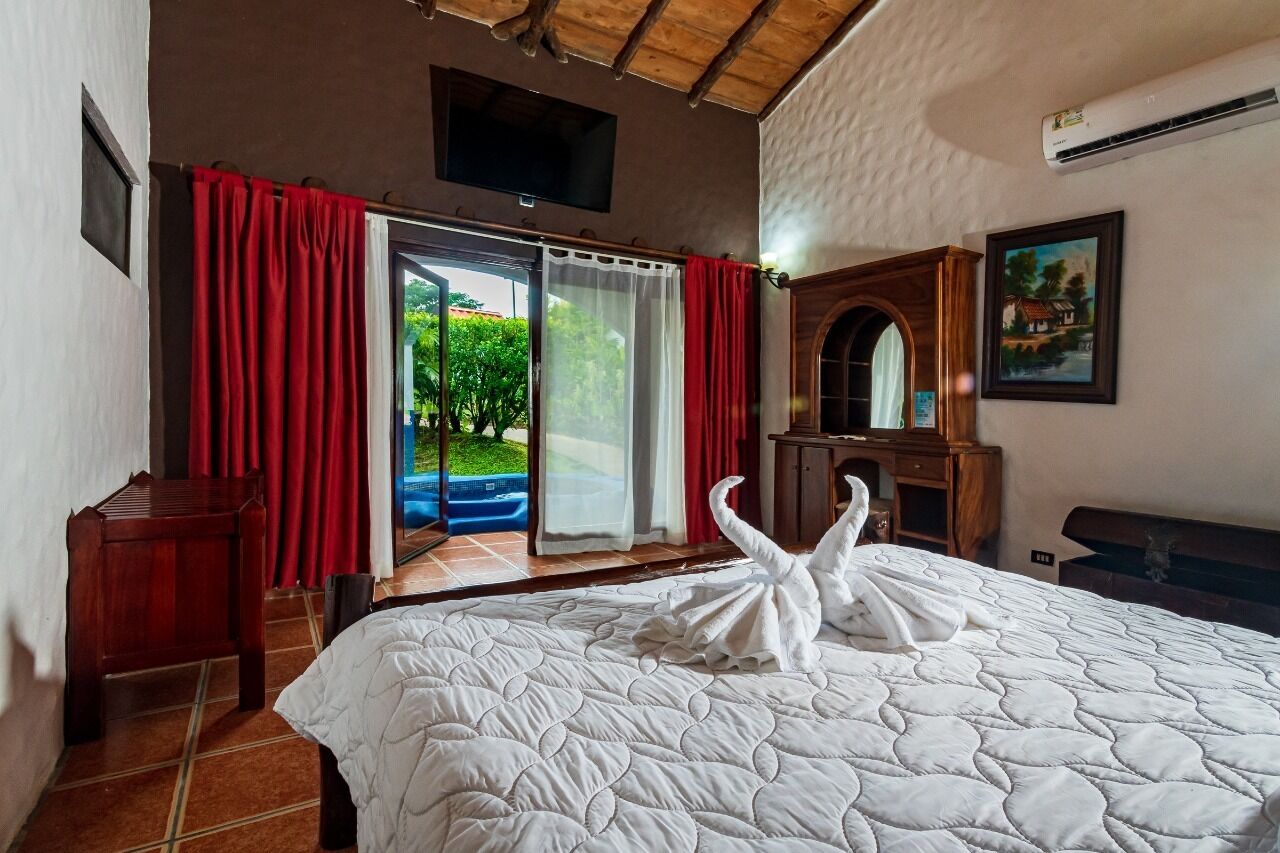 A hotel room with a bed, a wooden nightstand, a TV, a painting, and a view of greenery through the sliding glass door.
