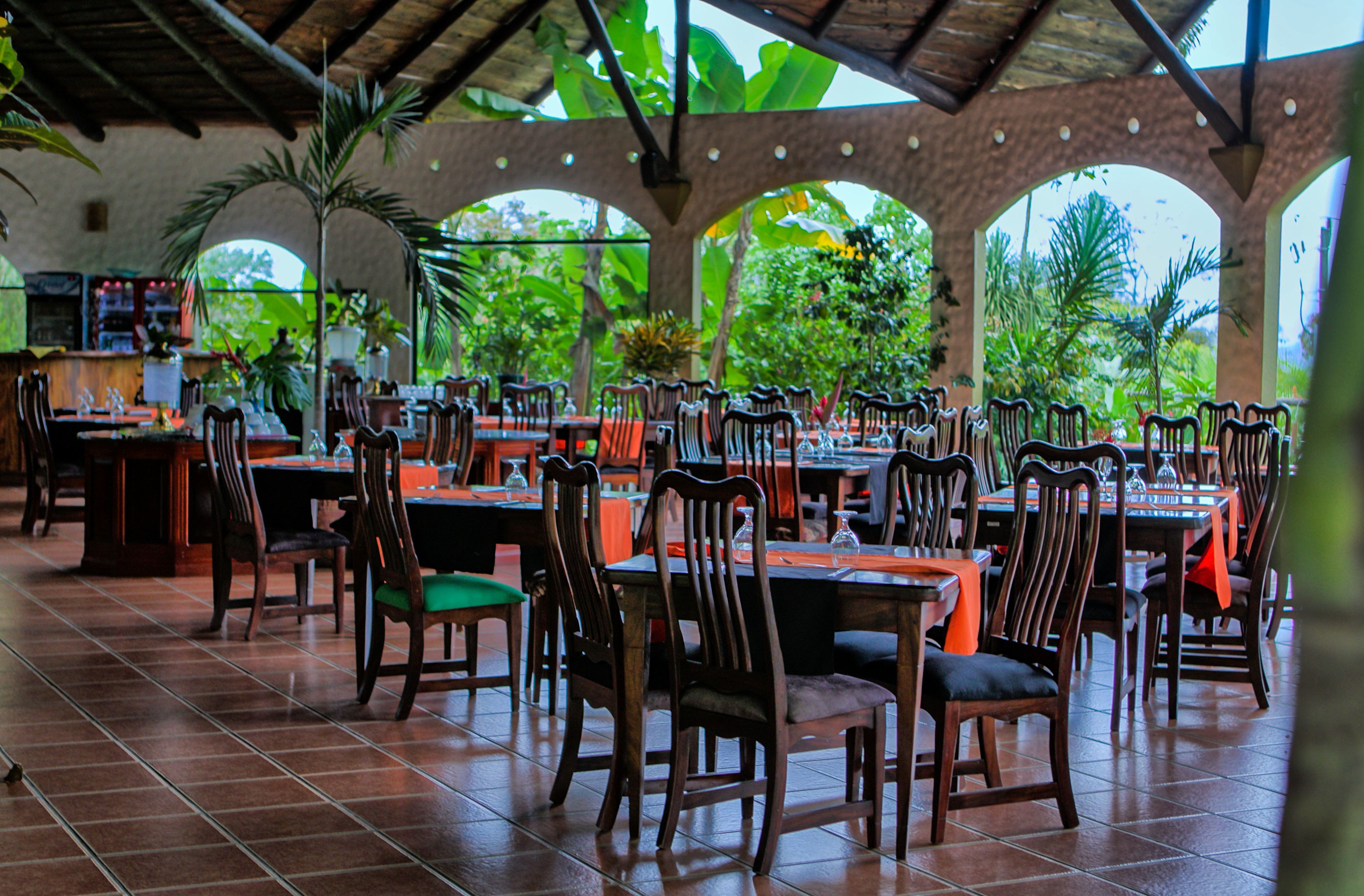 Un restaurante con varias mesas y sillas, rodeado de vegetación y un suelo de baldosas.