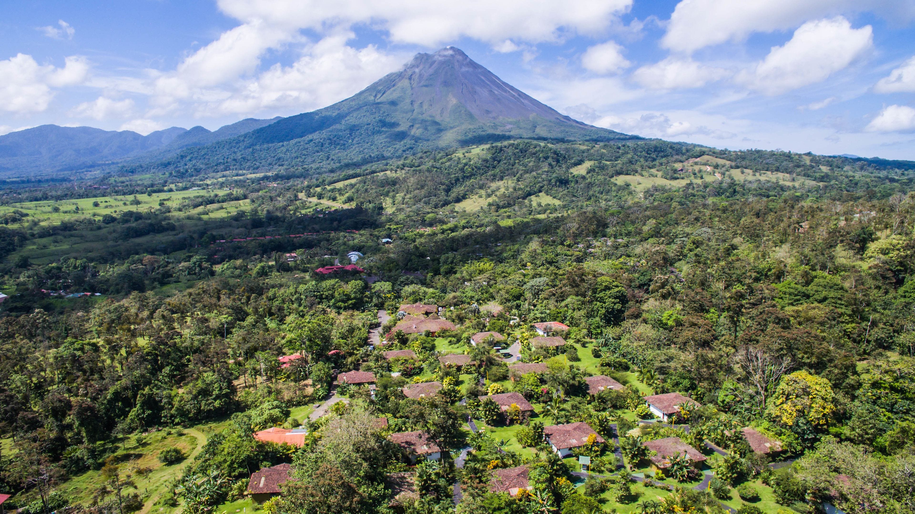 A mountainous landscape with a prominent volcano in the background and a village with houses surrounded by greenery in the foreground.
