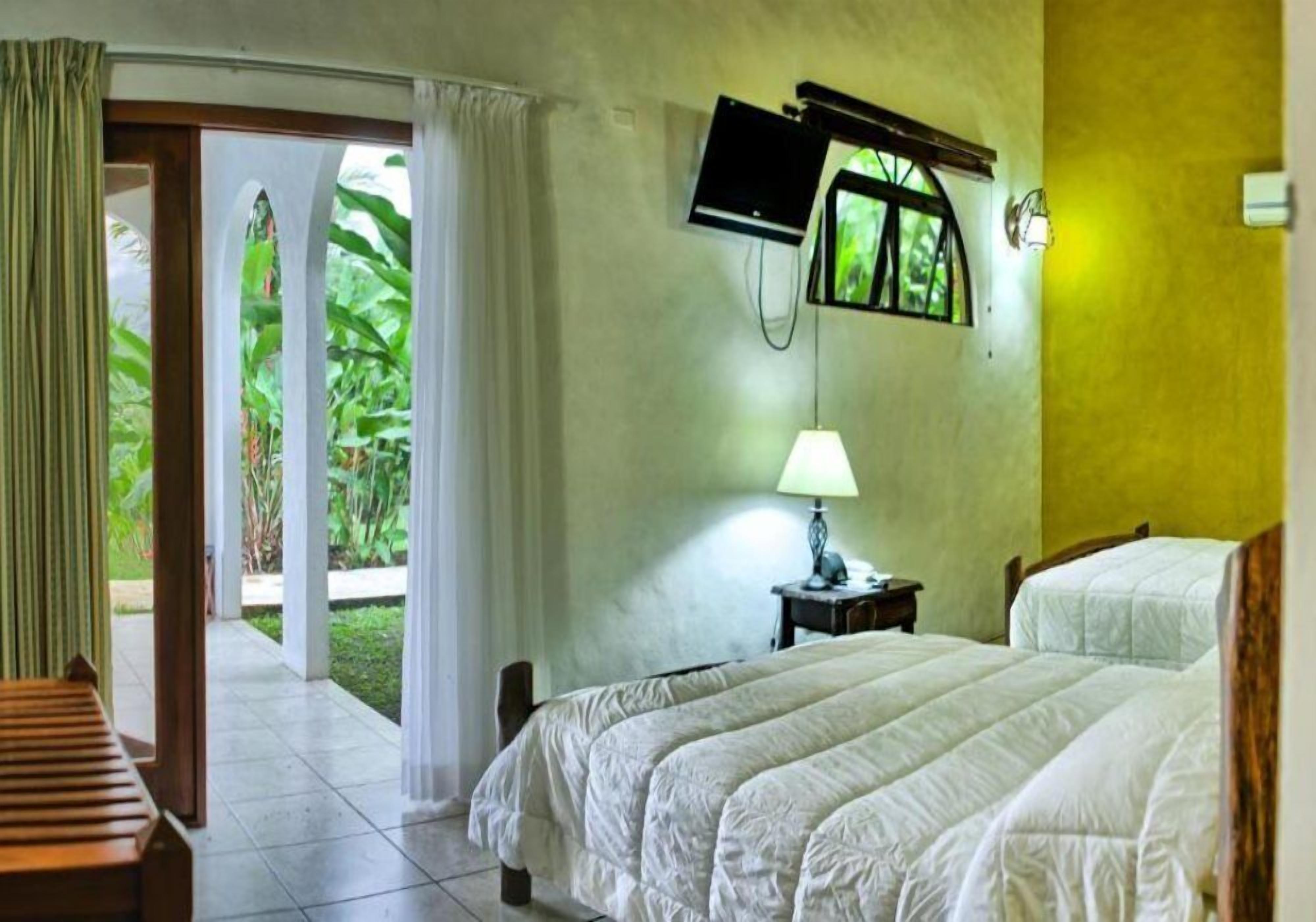 A hotel room with a bed, a TV mounted on the wall, a lamp, and a view of greenery outside.