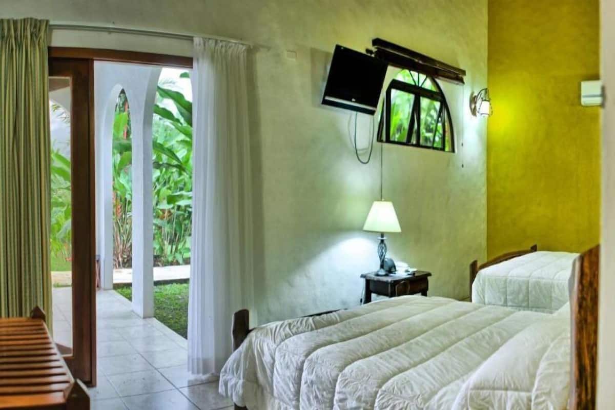 Une chambre d’hôtel avec un lit, un téléviseur fixé au mur, une lampe et une vue sur la verdure.