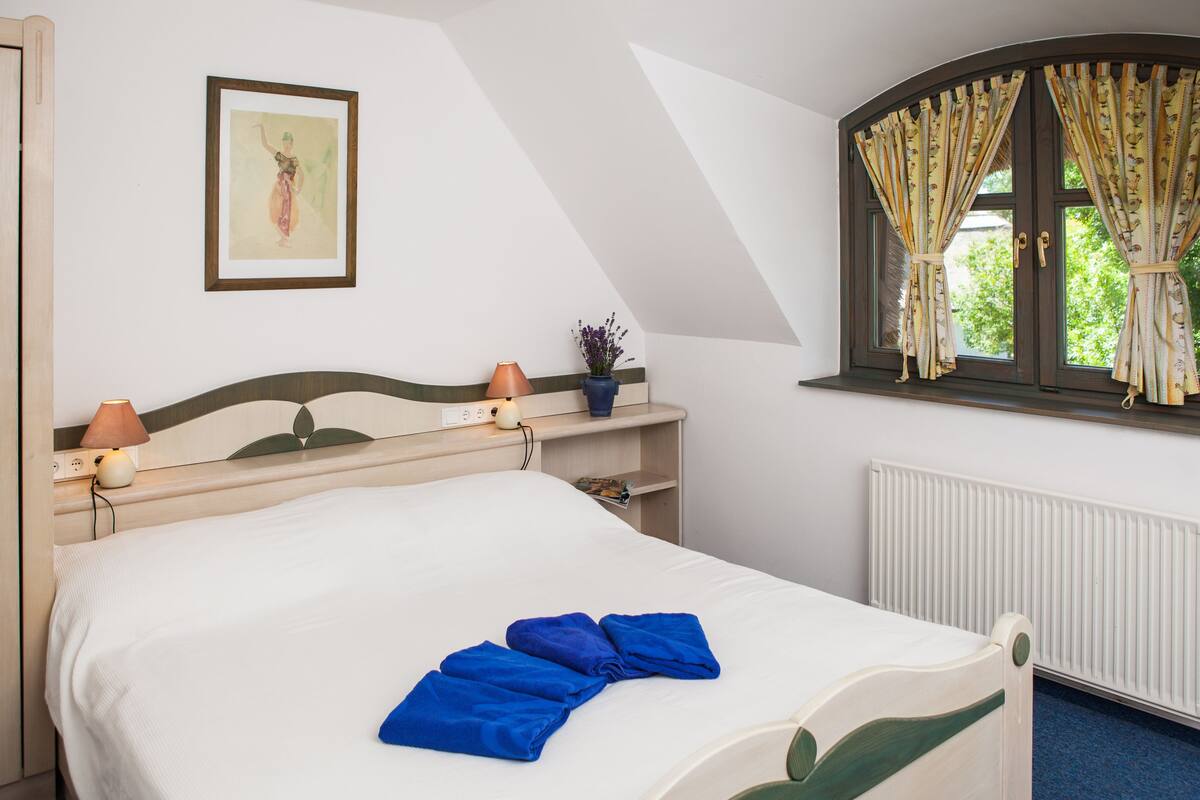 A hotel room with a bed, a lamp, a framed picture on the wall, a potted plant, and a window with curtains.