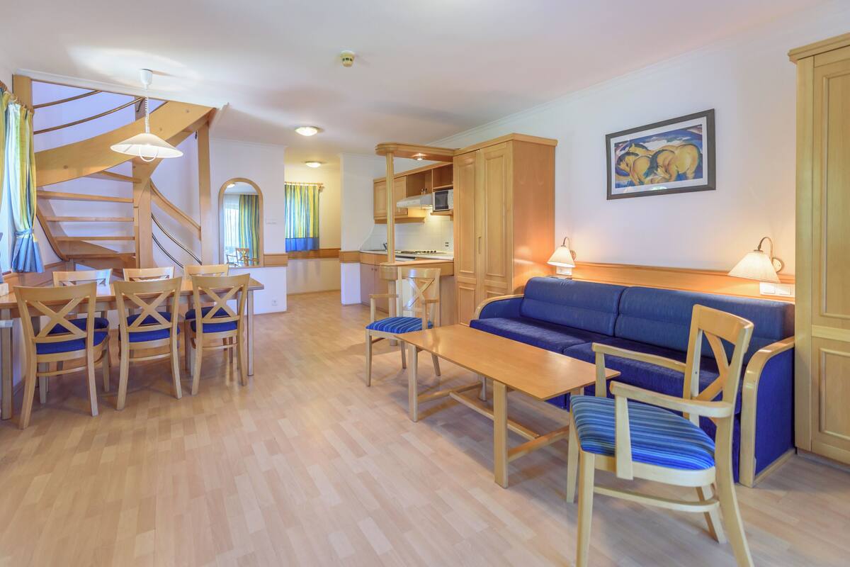A spacious living area with a wooden dining table, blue upholstered sofa, and matching chairs. A staircase with wooden railings leads to an upper level.