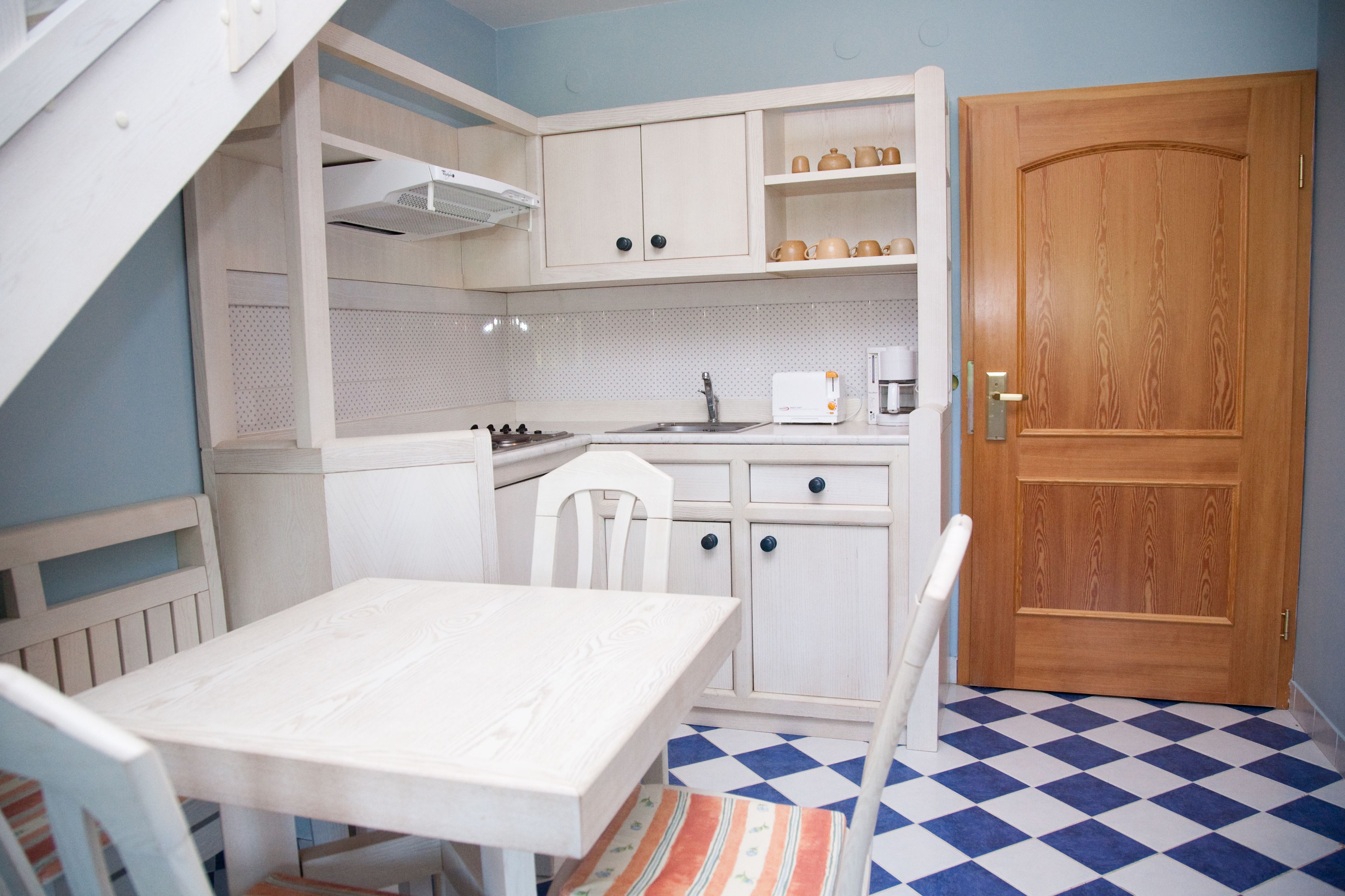 A compact kitchen with white cabinets, a stove, a sink, and a toaster. There is a wooden door and a checkered floor.