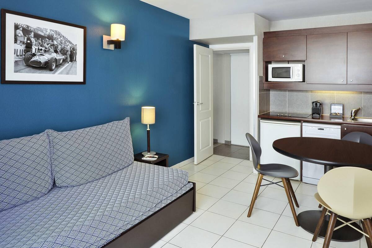 A modern room with a blue wall, a framed photo, a sofa, a bedside table with a lamp, a dining area with a table and chairs, and a kitchen with cabinets and appliances.