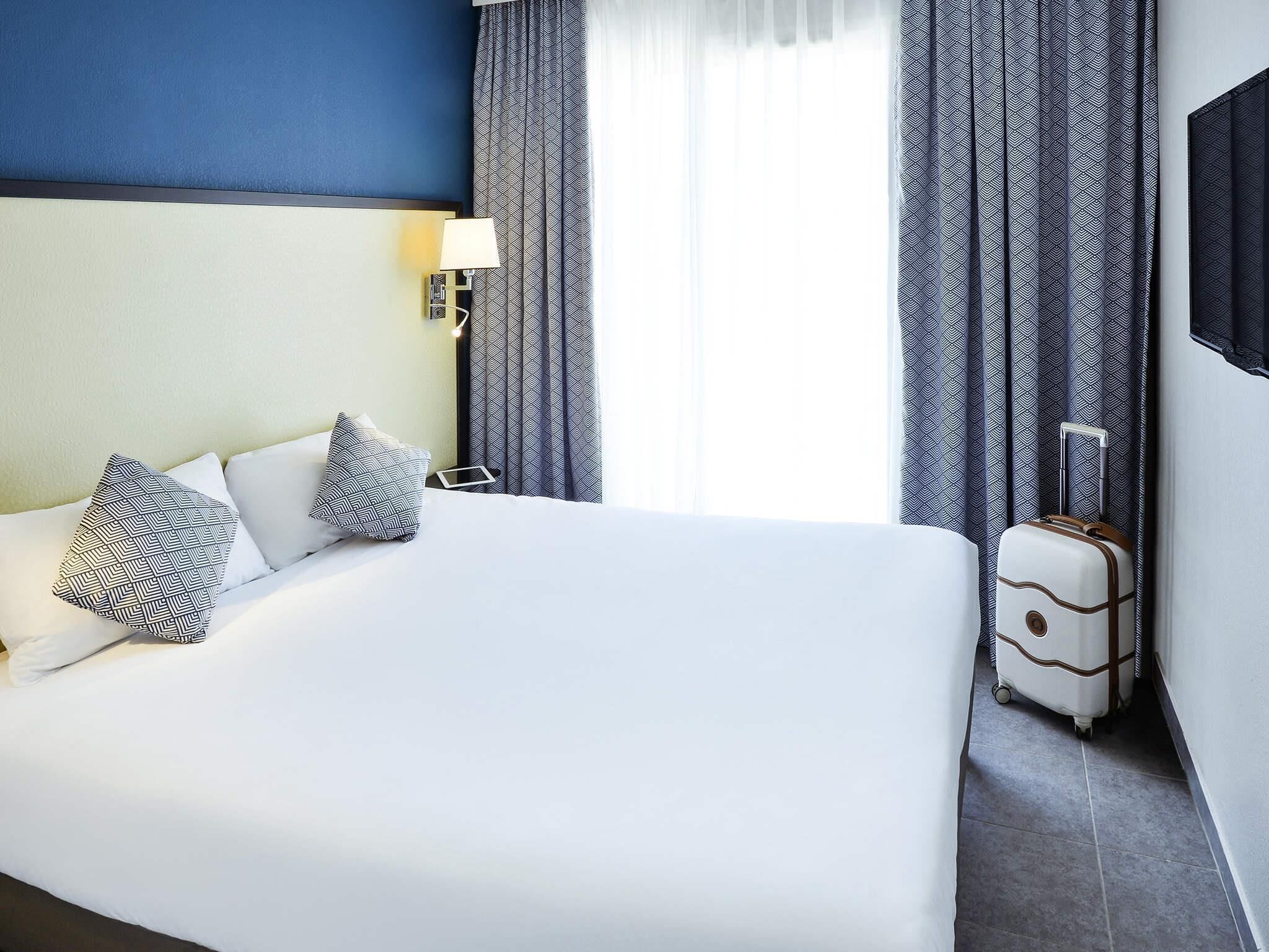 Una camera d'albergo con un letto grande, un comodino, una lampada, tende a motivi e una valigia.