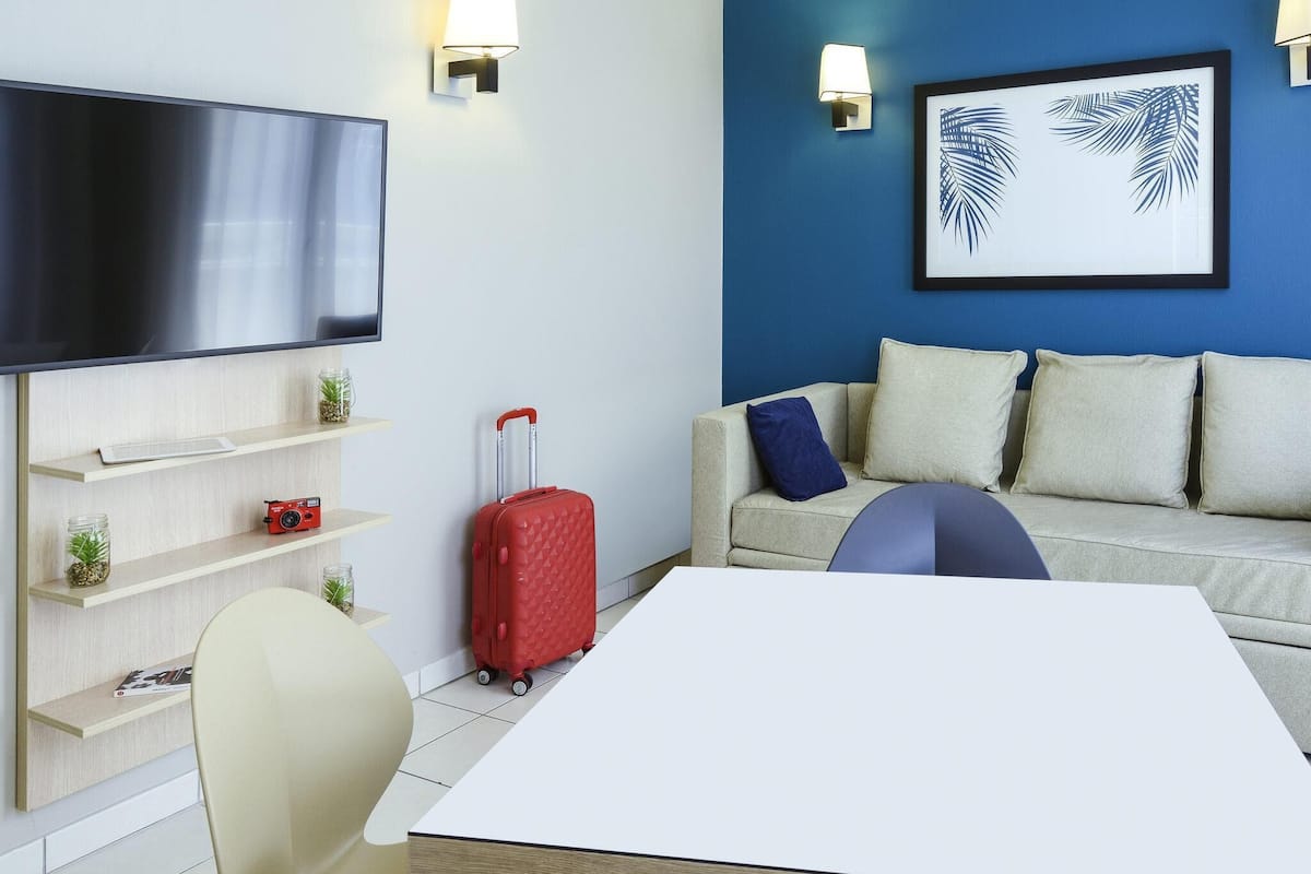 A modern hotel room with a flat-screen TV, a red suitcase, a sofa with cushions, and a framed artwork on the wall.