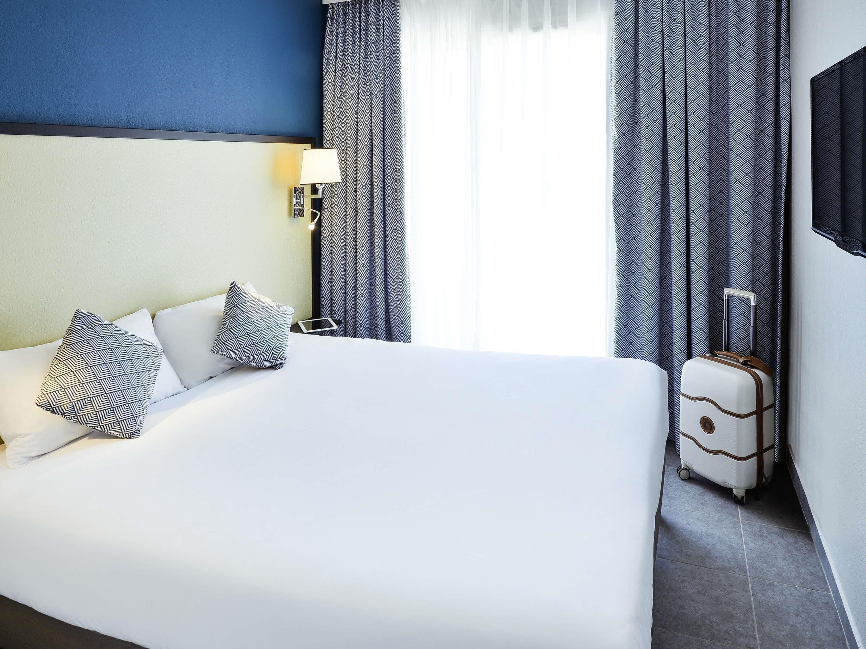 Hotellihuone, jossa on suuri sänky, yöpöytä, lamppu, kuviolliset verhot ja matkalaukku.