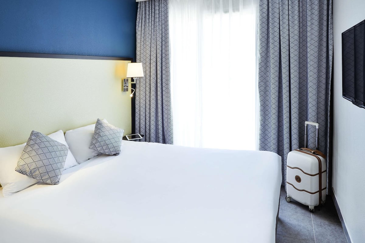Ein Hotelzimmer mit einem großen Bett, einem Nachttisch, einer Lampe, gemusterten Vorhängen und einem Koffer.