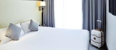 A hotel room with a large bed, a bedside table, a lamp, patterned curtains, and a suitcase.
