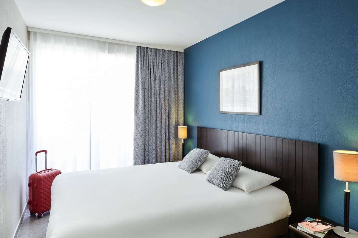Ein Hotelzimmer mit einem großen Bett, einem Nachttisch, einer Lampe, einem Koffer und einem Fenster mit Vorhängen.