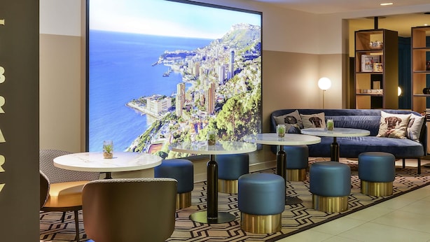 A modern lounge area with a large TV screen displaying a coastal cityscape, comfortable seating, and a patterned floor. - Aparthotel Adagio Monaco Monte Cristo (Beausoleil)