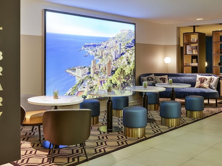 A modern lounge area with a large TV screen displaying a coastal cityscape, comfortable seating, and a patterned floor.. Aparthotel Adagio Monaco Monte Cristo