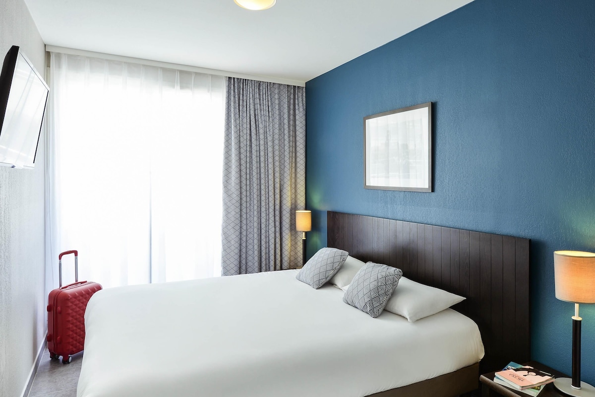 A hotel room with a large bed, a nightstand, a lamp, a suitcase, and a window with curtains.