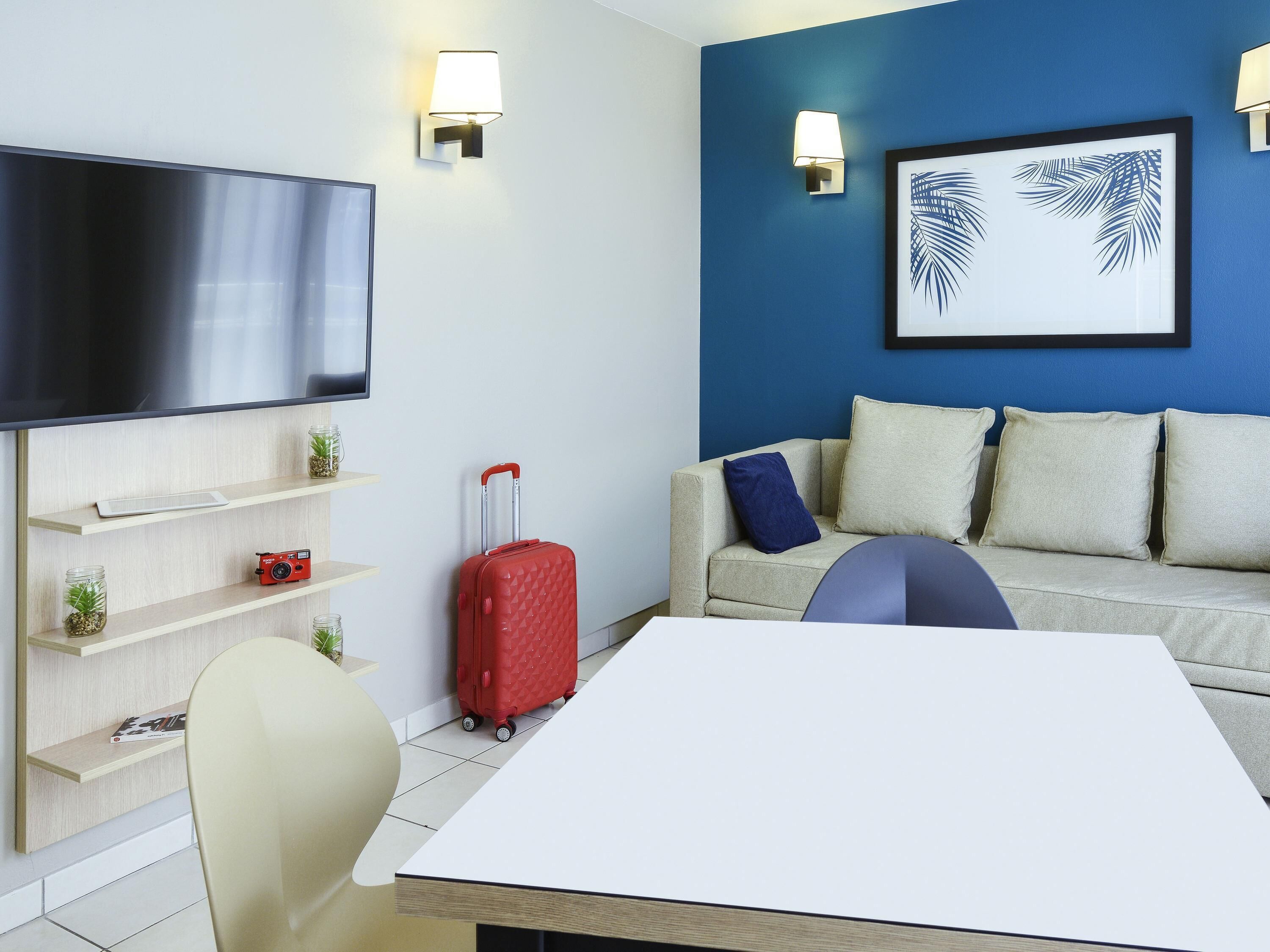 Ett modernt hotellrum med en platt-TV, en soffa med kuddar, en röd resväska, ett bord och väggmonterade lampor.
