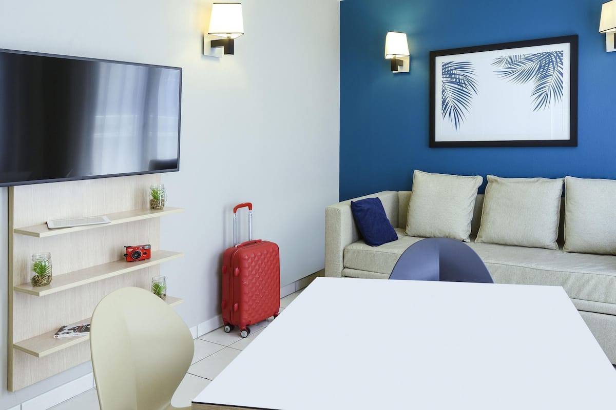 Ett modernt hotellrum med en platt-TV, en soffa med kuddar, en röd resväska, ett bord och väggmonterade lampor.