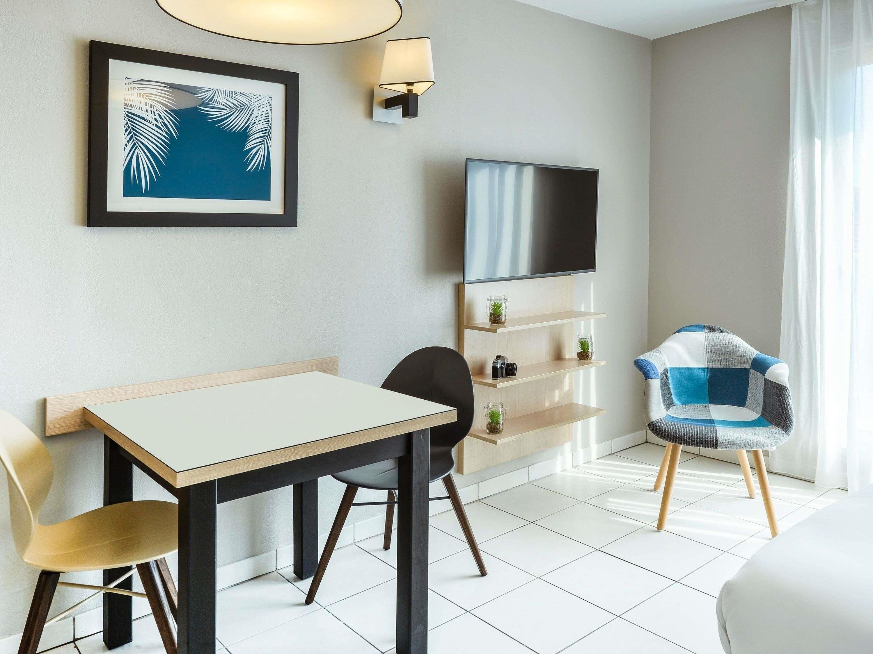 A hotel room with a bed, a desk with a chair, a television, a wall-mounted shelf with plants, and a framed picture.