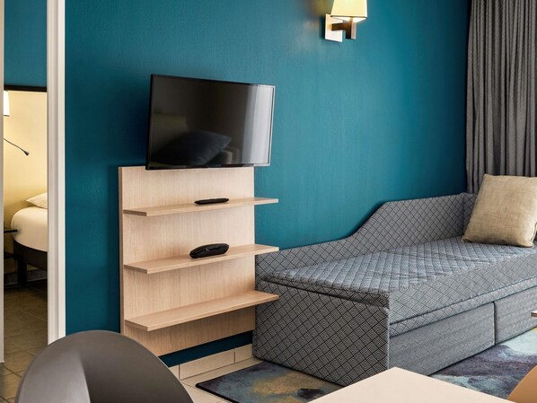 Habitación de hotel moderna con una pared verde azulado, un televisor de pantalla plana montado en un estante de madera, un sofá estampado y una mesita de noche con una lámpara.