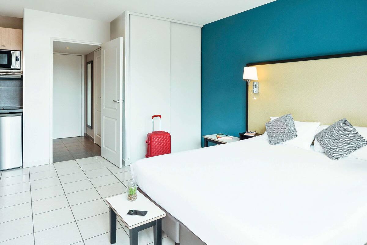 A hotel room with a bed, a nightstand, a red suitcase, a small table with a glass jar, and a microwave in the kitchen area.