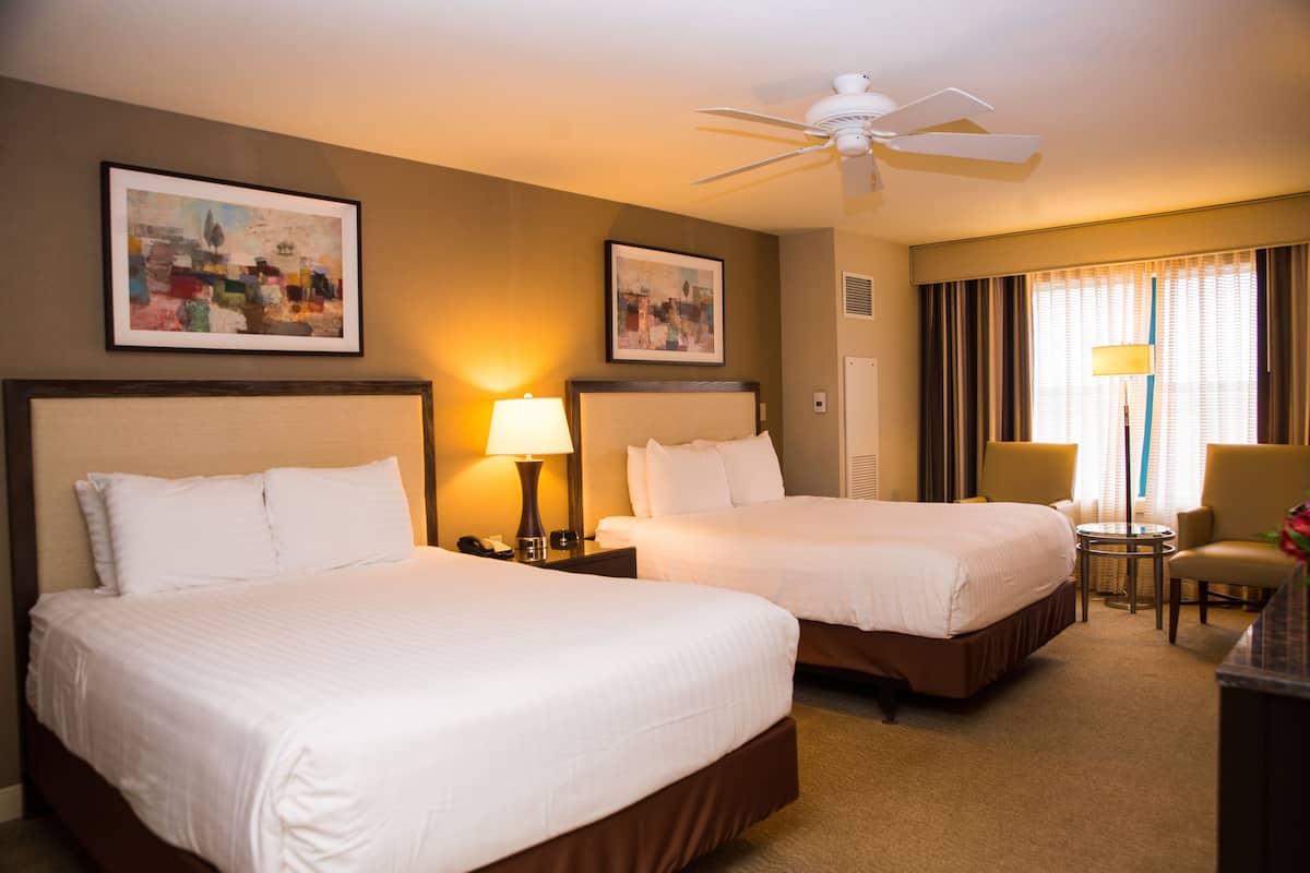 A hotel room with two beds, a ceiling fan, a desk with a chair, and artwork on the walls.