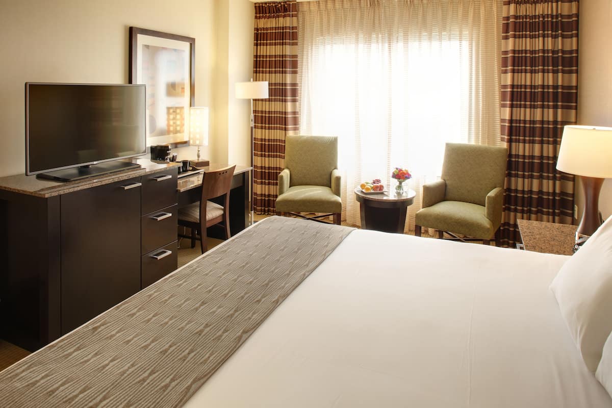 A hotel room with a bed, a desk with a television, two armchairs, a small table with a vase of flowers, and curtains.