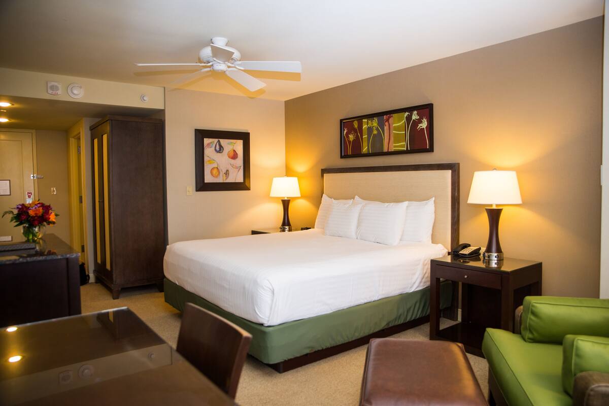 A hotel room with a bed, two bedside tables with lamps, a desk, a chair, a green sofa, and a ceiling fan.