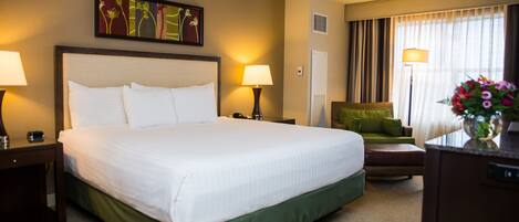 A hotel room with a large bed, two bedside tables with lamps, a green sofa, a desk with a lamp, and a window with curtains.
