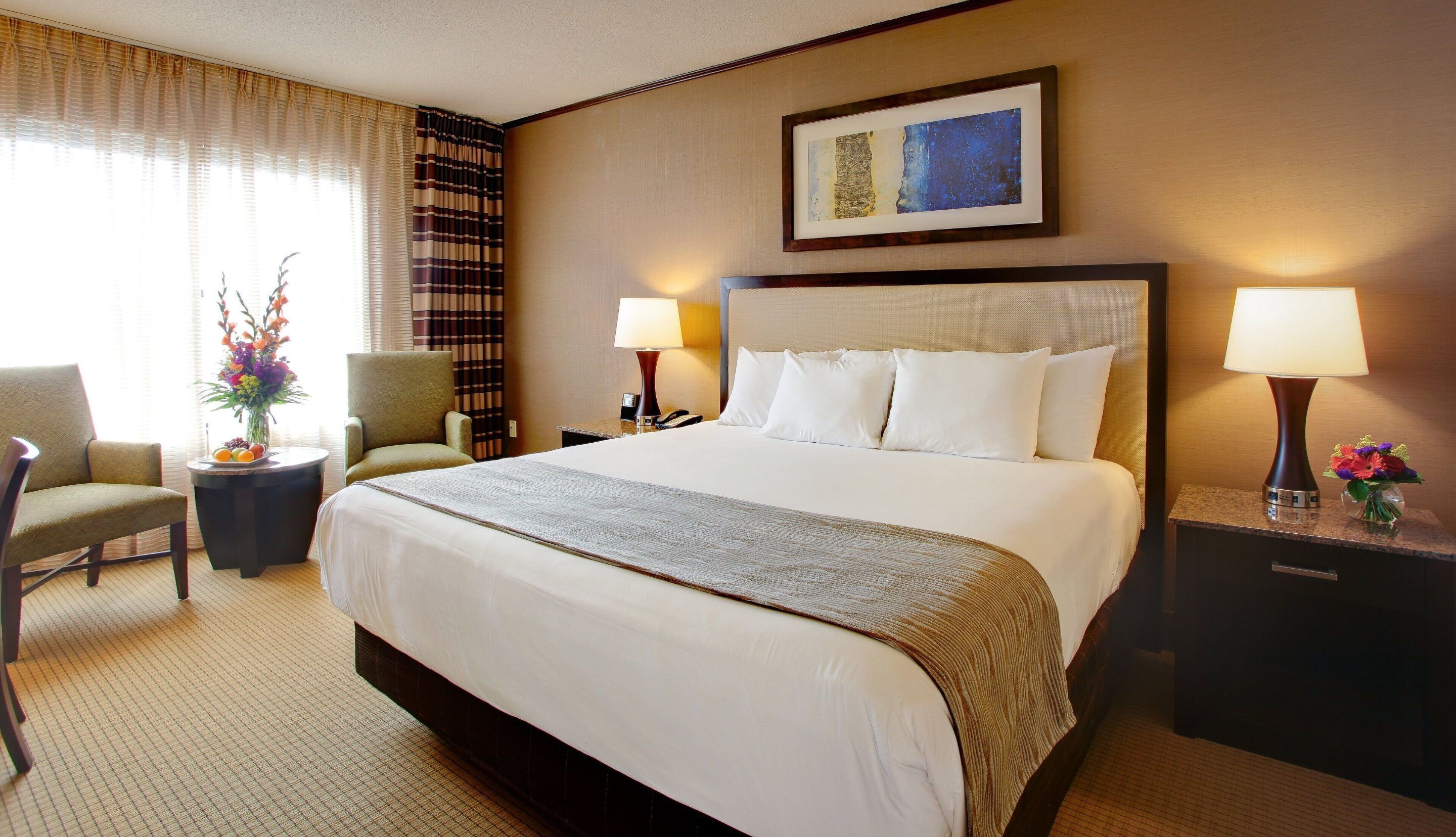 A hotel room with a large bed, two bedside lamps, a chair, a small table with a vase of flowers, and a framed picture on the wall.
