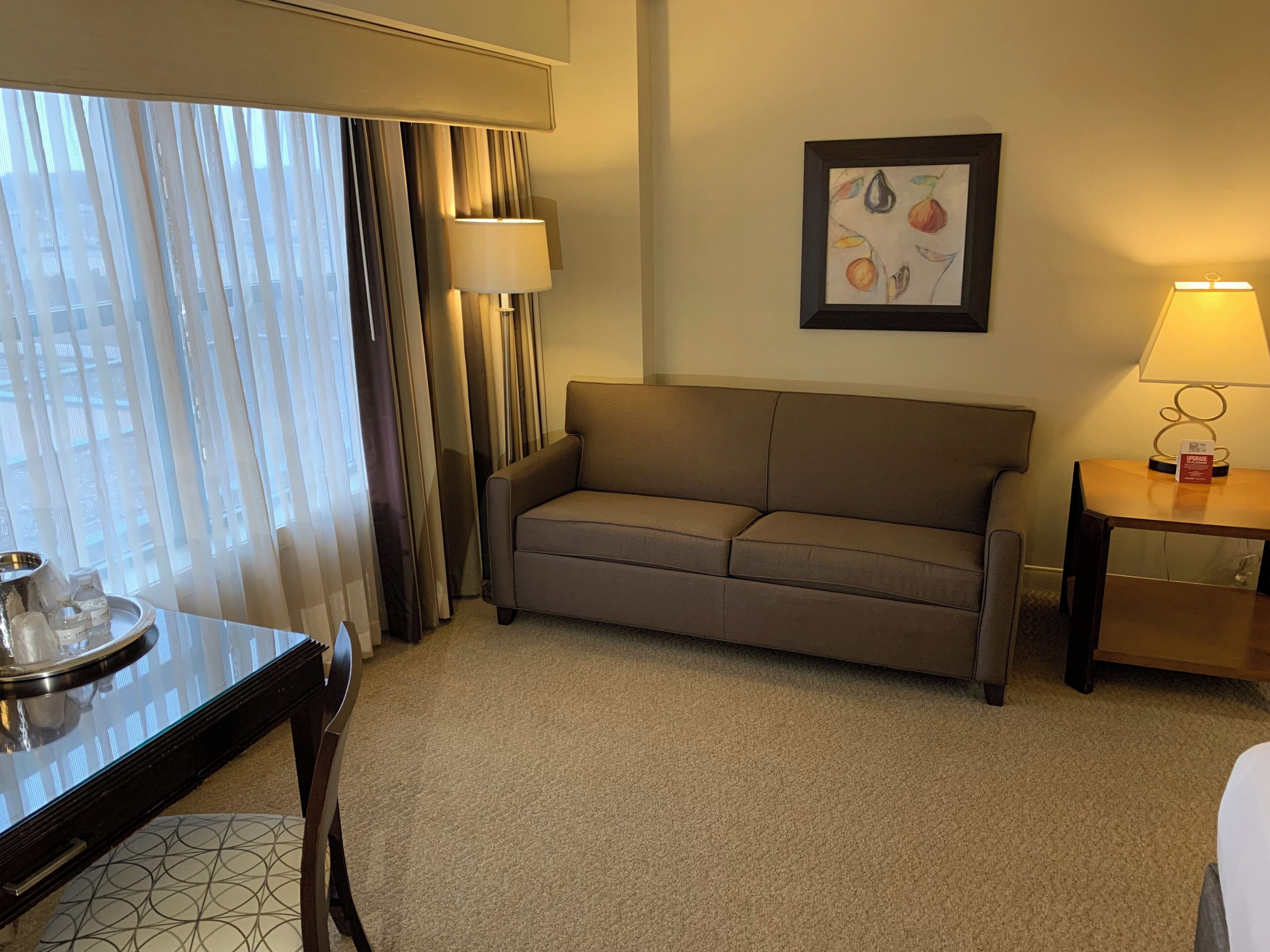 Habitación de hotel con un sofá color beige, una mesita con lámpara, una obra de arte enmarcada en la pared y una ventana con cortinas.