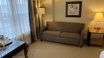 Une chambre d’hôtel avec un canapé beige, une petite table avec une lampe, une œuvre d’art encadrée au mur et une fenêtre avec des rideaux.