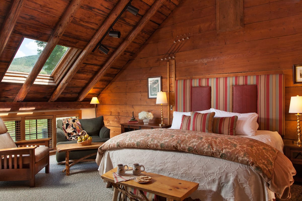 A cozy room with a wooden ceiling, a large bed with striped pillows, a small table with a basket and mugs, and a view of greenery through skylights.