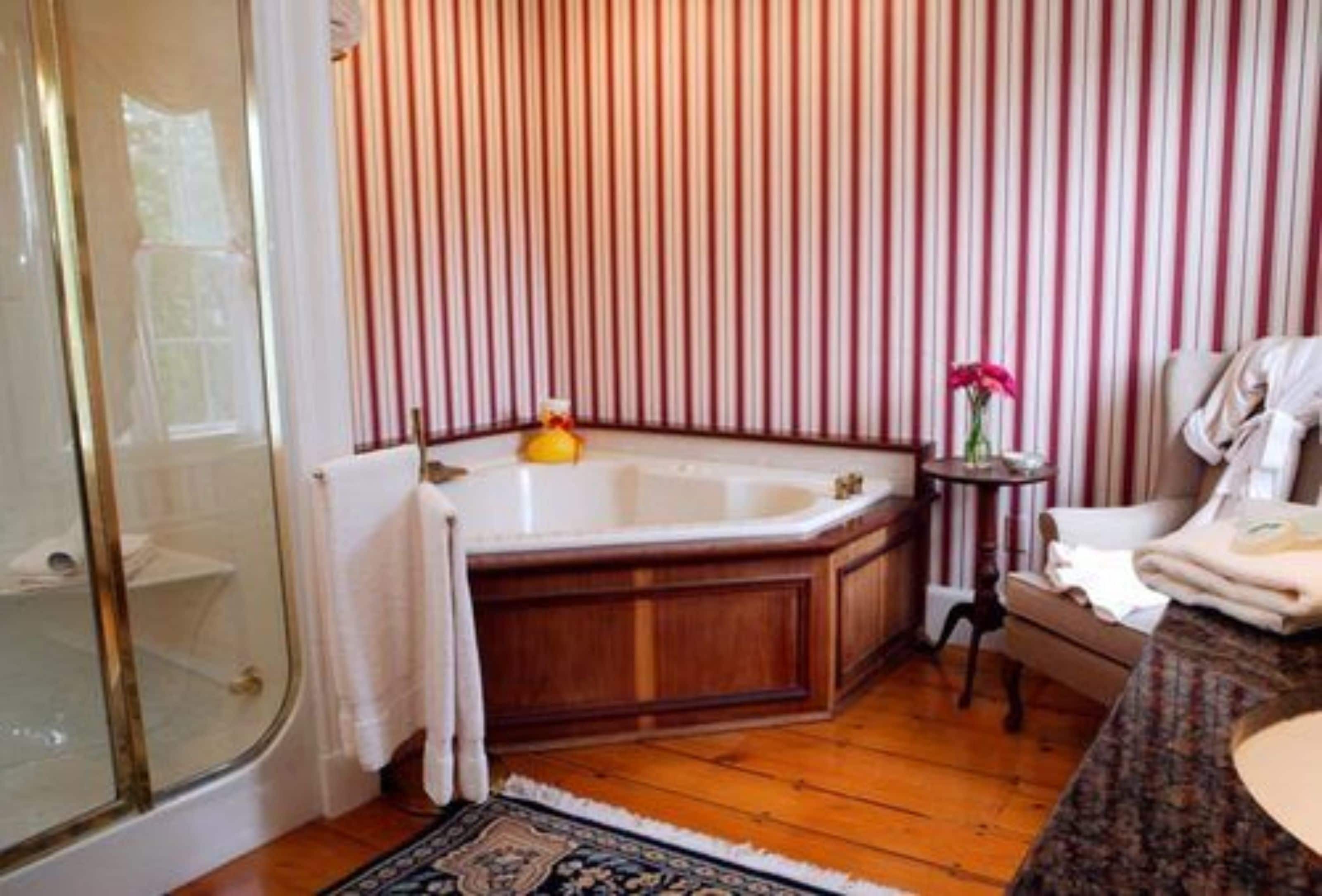 A bathroom with a bathtub, a glass shower enclosure, striped wallpaper, and a small table with a vase.
