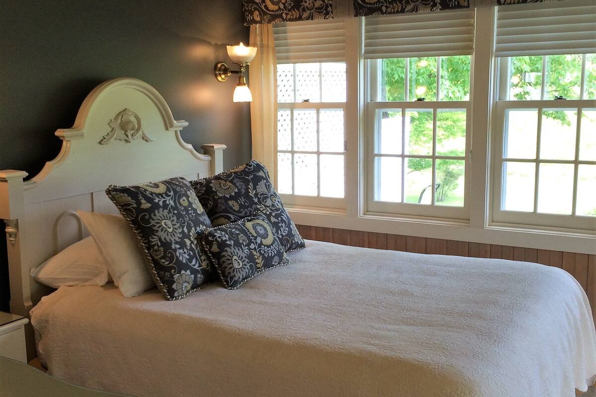 A bedroom with a large bed, a nightstand, a lamp, and a window with a view of greenery.