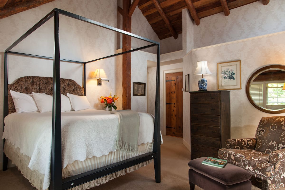 A four-poster bed with a white bedspread and pillows, a vase with flowers, a wooden chair, and a round mirror on the wall.