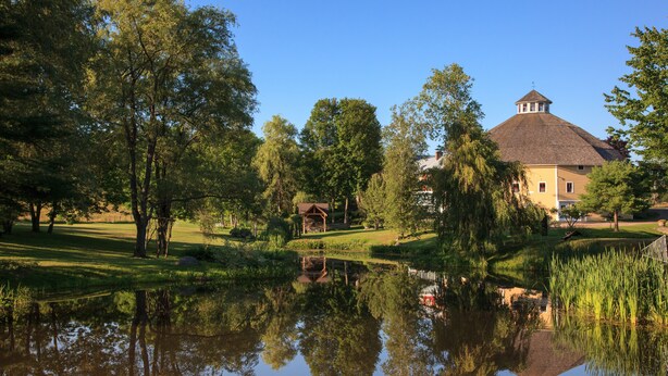 A serene lakeside setting with a traditional building featuring a tower, surrounded by lush greenery and a calm pond reflecting the structure. - The Inn at Round Barn Farm (Waitsfield)