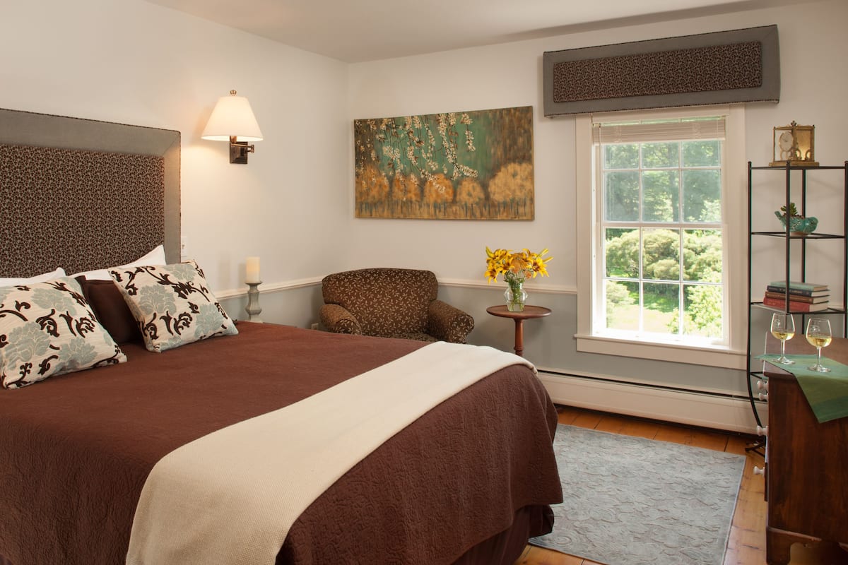 A bedroom with a large bed, a chair, a small table with flowers, a bookshelf, and a window with a view of greenery.