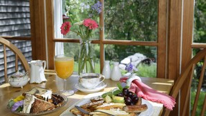 A breakfast table with a variety of food items, a vase with flowers, and a view of greenery through a window. - The Inn at Round Barn Farm (Waitsfield)