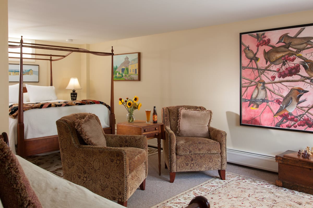 A hotel room with a four-poster bed, two patterned armchairs, a small table with a vase of sunflowers, a painting of birds, and a wooden nightstand.