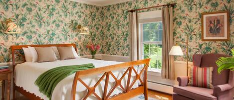 A bedroom with a wooden bed, floral wallpaper, a window with curtains, a chair, and a lamp.