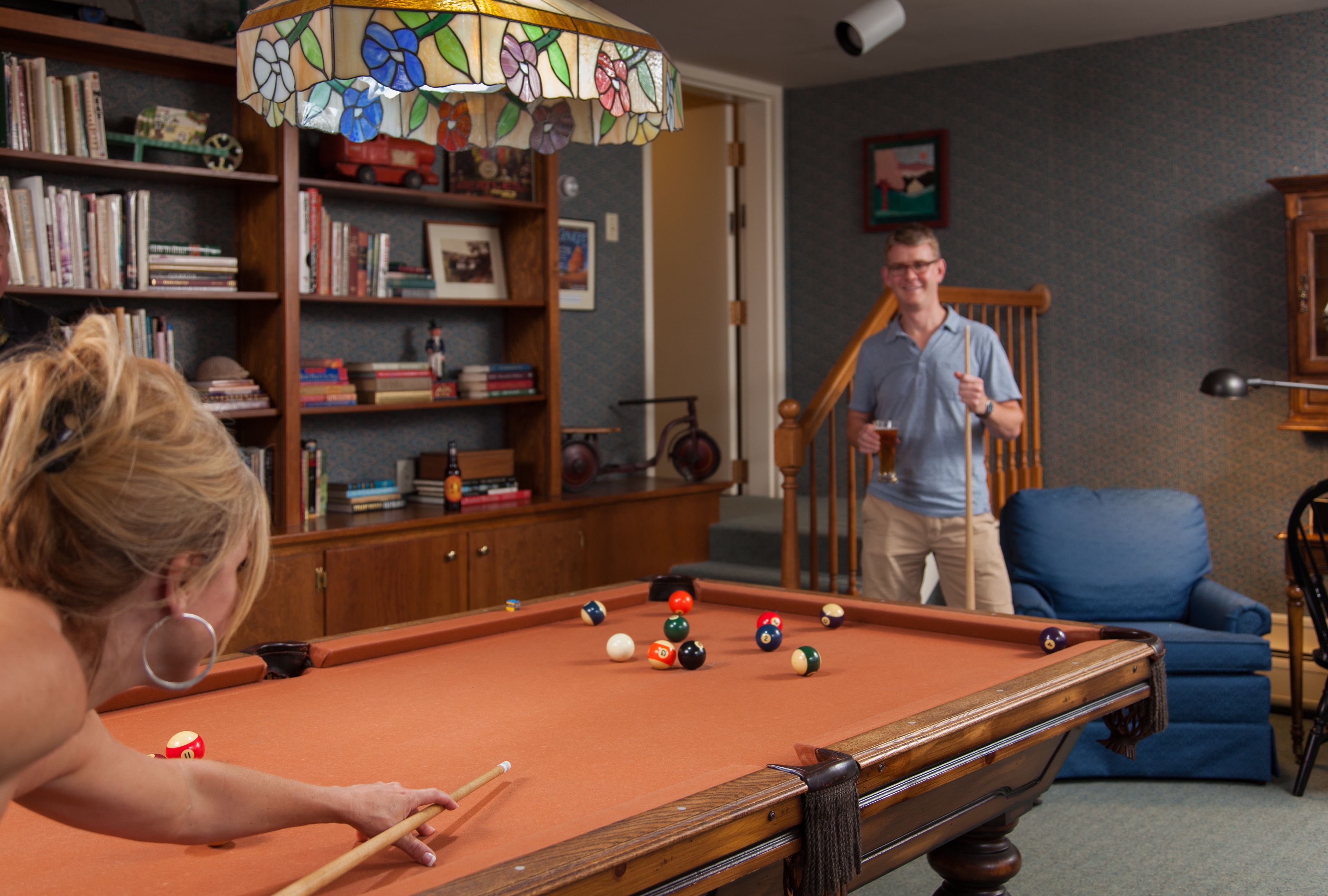 A pool table in a room with a bookshelf, a blue armchair, and a staircase.