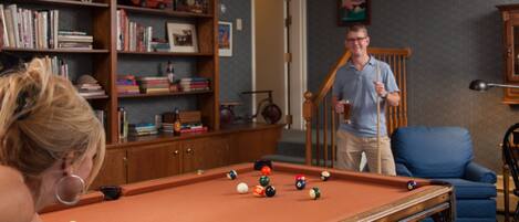 A pool table in a room with a bookshelf, a blue armchair, and a staircase.