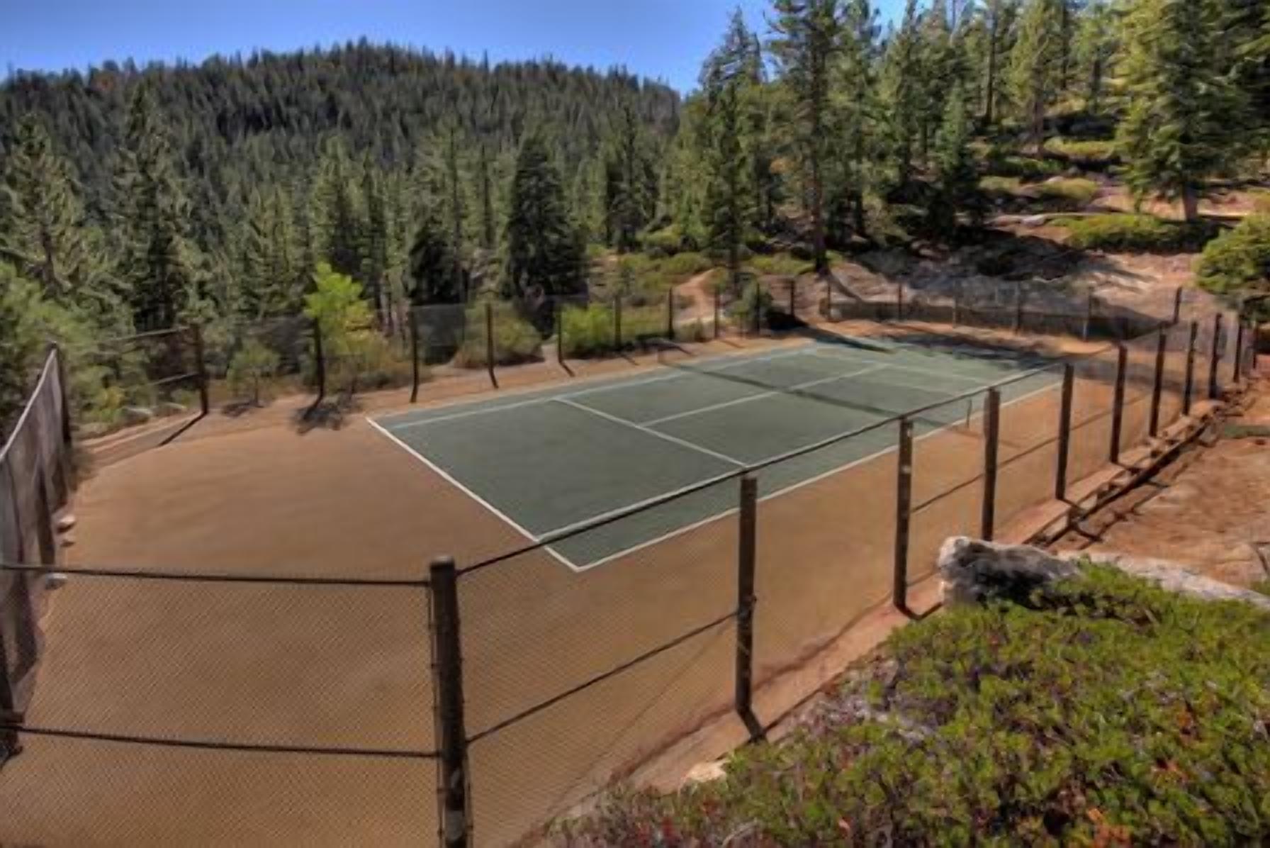 A tennis court surrounded by a fence, with a forested hillside in the background.