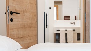 A bedroom with a wooden door, a bed with white bedding, a white bedside table, and a bathroom with a mirror, towel rack, and wooden chair. - CHRIZZ Rooms (Graz)