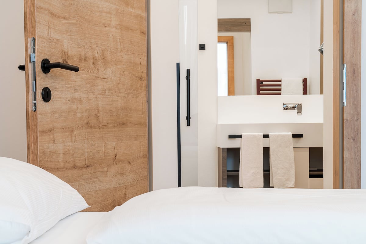 Ein Schlafzimmer mit Holztür, einem Bett mit weißer Bettwäsche, einem weißen Nachttisch sowie einem Badezimmer mit Spiegel, Handtuchhalter und Holzstuhl.